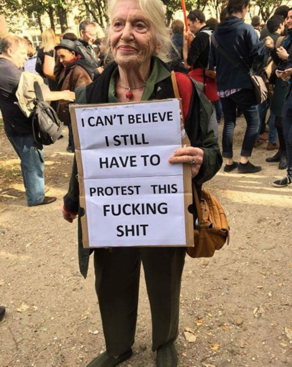 Charismatic middle aged woman at a protest with handmade sign that says 'I CANT BELIEVE I STILL HAVE TO PROTEST THIS FUCKING SHIT'